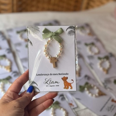 Lembrança de batizado com pulseira de contas peroladas, cruz dourada e laço verde num cartão branco decorado