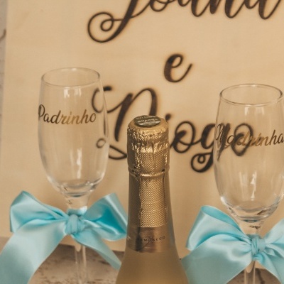 Duas taças de champanhe decoradas com laços azuis e uma garrafa de champanhe em frente a uma caixa bege com texto em português