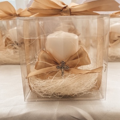 Vela branca decorativa com laço dourado e cruz dentro de caixa transparente