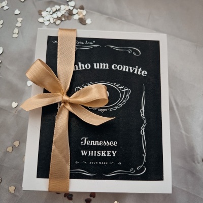 Cartão convite com fita dourada e texto Tennessee Whiskey sobre tecido cinza com confetes em formato de coração.