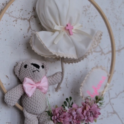 Coroa decorativa com ursinho de croché, flores cor-de-rosa e manta branca com cruz cor-de-rosa