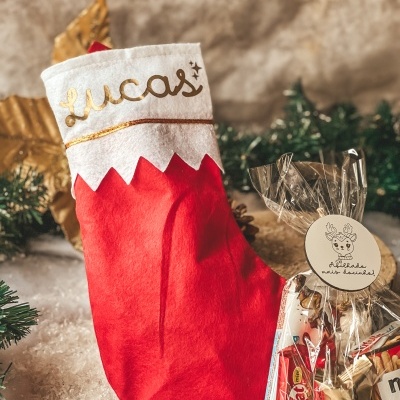Meia de Natal vermelha com nome Lucas em dourado e saco de chocolates ao lado, cenário de neve artificial e decoração natalícia.