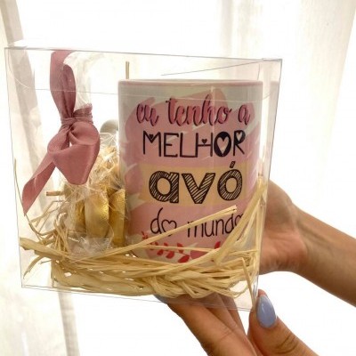 Caneca com texto, saco dourado e laço rosa dentro de embalagem transparente, segurada por mãos com unhas azuis.