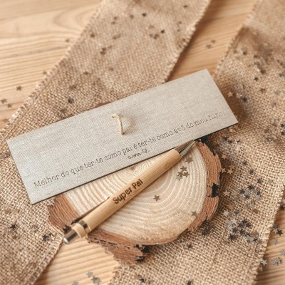 Caneta de madeira clara com texto Super Pai e cartão com texto em português sobre superfície de madeira e tecido rústico