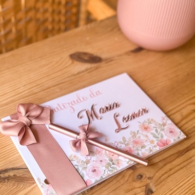 Livro de batizado com decoração rosa e dourada e caneta sobre mesa de madeira