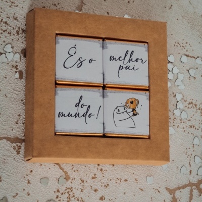 Caixa com quatro chocolates embrulhados em papel branco com texto e desenho, sobre uma superfície com pequenos corações brancos espalhados.