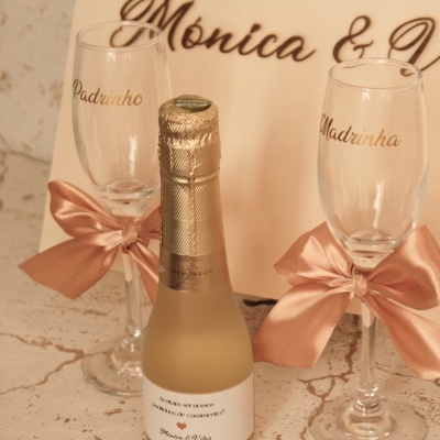 Duas flutes de champanhe com laços cor-de-rosa e texto 'Padrinho' e 'Madrinha', pequena garrafa de champanhe e fundo com texto 'Mónica & Vítor'