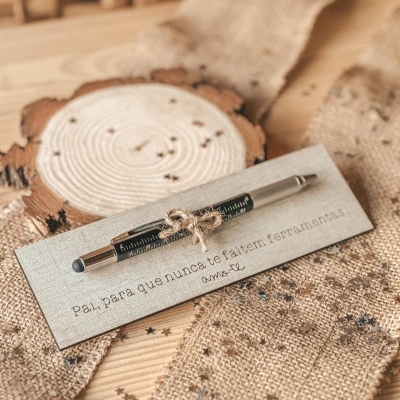 Caneta preta com régua impressa em cartão cinzento com mensagem em português sobre superfície de madeira e juta