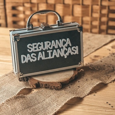 Mala preta pequena com texto branco SEGURANÇA DAS ALIANÇAS! sobre mesa de madeira com tecido rústico
