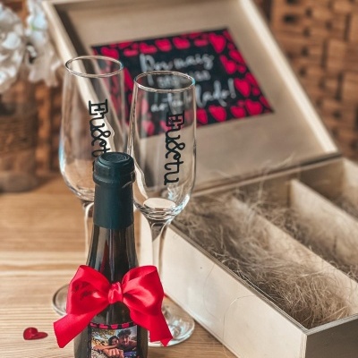 Garrafa de champanhe pequena com laço vermelho e duas taças com texto junto a caixa de madeira aberta.