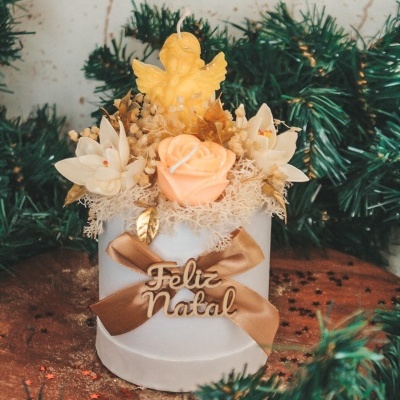 Vela branca decorativa de Natal com laço castanho e placa 'Feliz Natal', flores falsas e anjo amarelo no topo.