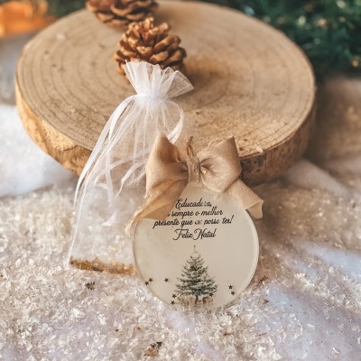 Enfeite de Natal redondo branco com laço bege e texto em cima de neve artificial com cones de pinheiro.