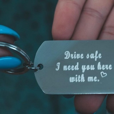 Chaveiro metálico prateado com gravação branca e coração, segurado por mão com unhas azuis.