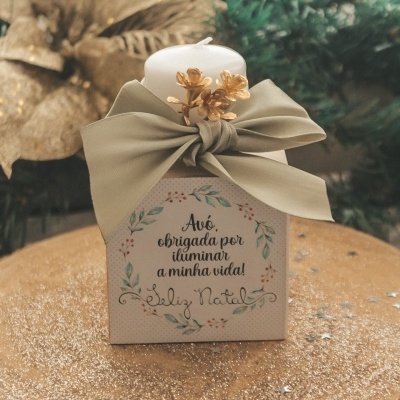 vela branca decorativa com laço verde e flores douradas em caixa com texto de Natal