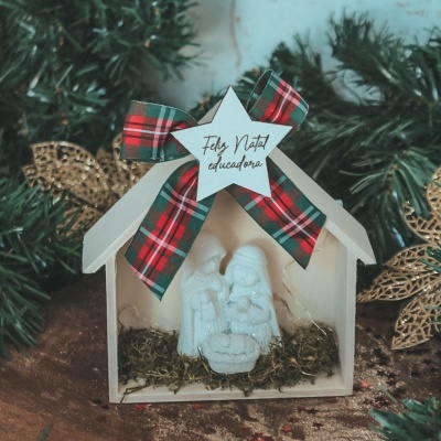 Presépio pequeno branco com figuras brancas e laço xadrez numa mesa com ramos verdes e folhas douradas.