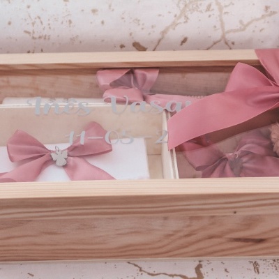 Caixa de madeira com tampa de vidro e laço rosa, contendo tecido branco com laço rosa e flores artificiais.