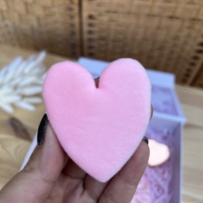 Esponja rosa em forma de coração segurada por uma mão, com uma caixa branca desfocada ao fundo