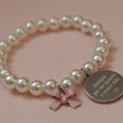 Pulseira de contas pérola com pendente redondo e laço cor-de-rosa, sobre fundo rosa claro.