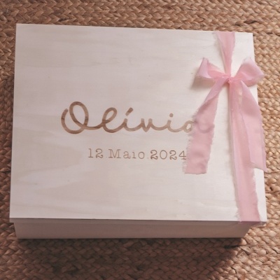 Estojo de madeira clara gravado com nome e data, laço rosa, sobre superfície de tecido texturado