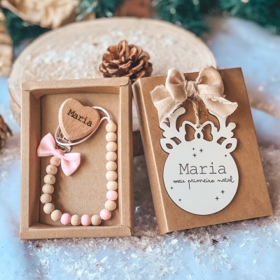 Prendedor de chupeta em contas de madeira naturais e cor-de-rosa com coração de madeira gravado Maria, em caixa de papel kraft decorada com placa branca de rena e laço.