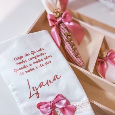Pano branco bordado com texto e laço rosa, e vela decorativa com laço rosa numa caixa de madeira.