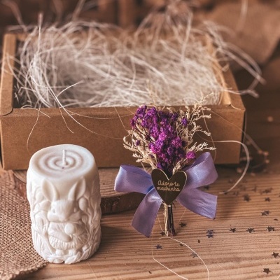 Vela decorativa branca ao lado de ramo de flores secas com laço lilás e placa coração e caixa de cartão