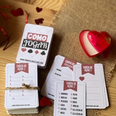 Baralho de cartas com instruções de jogo em português e decoração de corações vermelhos sobre mesa de madeira e tecido