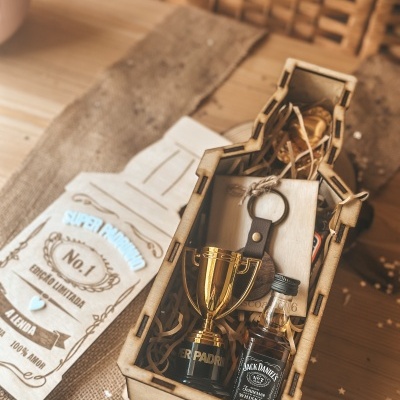 Cesta de madeira com troféu, mini garrafa de whisky e outros itens, sobre mesa com tecido rústico e papel decorativo.