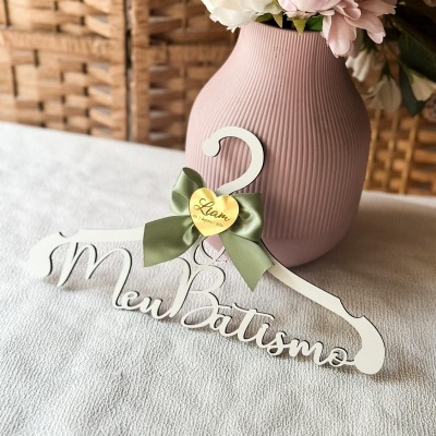 Cabide branco decorativo com texto Meu Batismo e laço verde ao lado de jarro rosa com flores sobre tecido branco