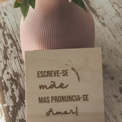 Placa de madeira clara com frase gravada e vaso cor-de-rosa com flores sobre mesa de madeira desgastada