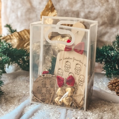 Saco presente transparente com objetos decorativos de madeira com desenhos e laços vermelhos em ambiente com neve falsa e decoração festiva