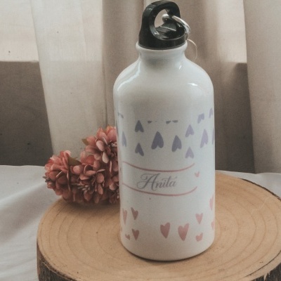 Garrafa de metal branca com corações coloridos e tampa preta sobre base de madeira com flores atrás