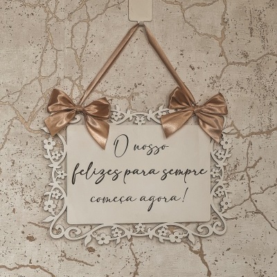 Placa decorativa branca com texto pendurada por fita com laços numa parede texturizada