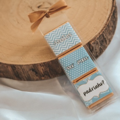 Embalagem transparente com chocolates decorados e texto ser meu padrinho, em fundo branco e base de madeira