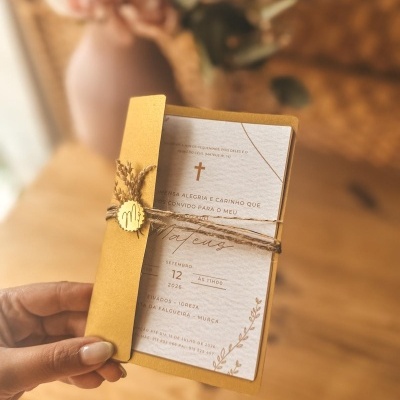 Convite em papel branco texturizado com envelope amarelo e decoração rústica em fundo interior desfocado.