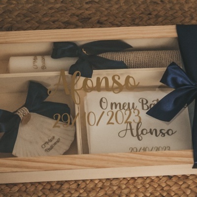 Caixa de presente em madeira clara com itens de batismo, incluindo laços e tecido bordado em azul escuro.