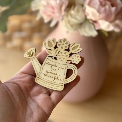 Ramo decorativo em madeira em forma de regador com flores e texto gravado, mão a segurá-lo, vaso cor-de-rosa com flores ao fundo.