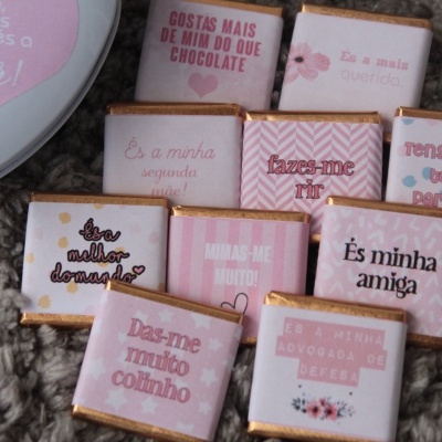 Chocolates pequenos embrulhados em papéis rosa, brancos e dourados com mensagens escritas em português sobre tapete cinzento.