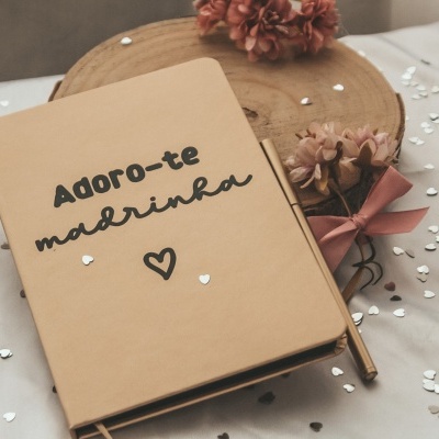 Caderno bege com texto 'Adoro-te madrinha' e flores num cenário branco.