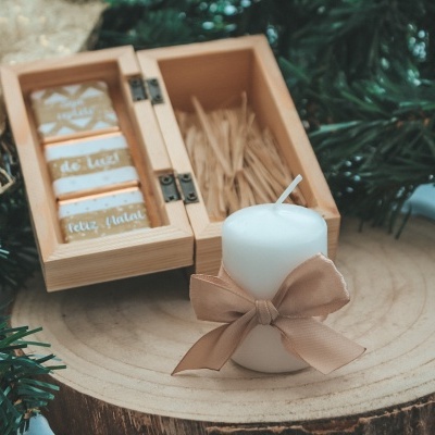 Vela branca com fita bege ao lado de caixa de madeira com palitos e etiquetas de madeira decorativas