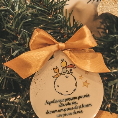 Ornamento de Natal redondo branco com laço dourado e texto em português pendurado numa árvore de Natal