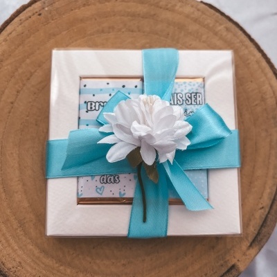 Caixa branca decorada com fita azul e flor branca sobre tronco de madeira