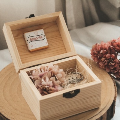 Caixa de madeira aberta com flores secas e bijuterias dentro, broche na tampa e flores ao lado.