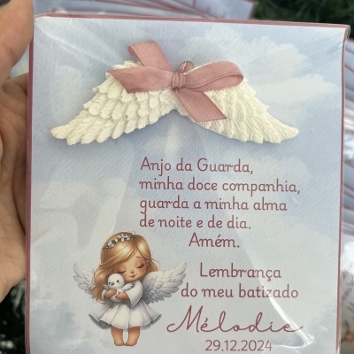 Embalagem com asas de anjo, fita rosa e texto de batizado em português