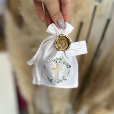 Saco branco com cruz dourada e coroa de flores, selo dourado e etiqueta, mão segurando, fundo com plantas secas