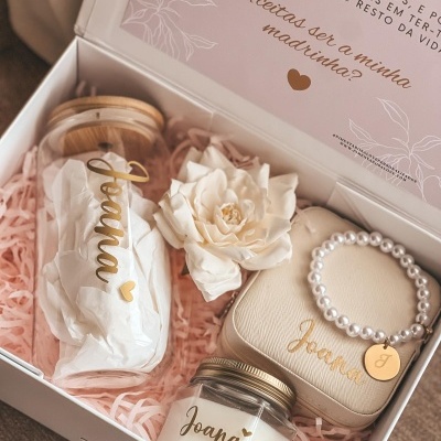 Caixa de oferta branca com itens personalizados com nome Joana em dourado, enchimento rosa, frasco, vela, estojo e pulseira.