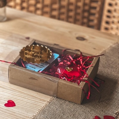 Caixa de presente em cartão com decoração vermelha e etiqueta dourada sobre mesa de madeira e passadeira de juta com corações