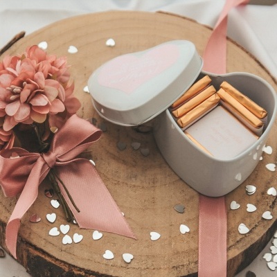 Caixa em forma de coração aberta com palitos, ramo de flores rosa e confetis de corações em base de madeira