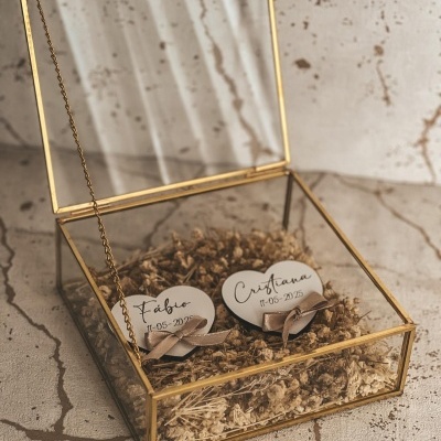 Caixa de vidro dourada aberta com corações de madeira com nomes sobre musgo seco