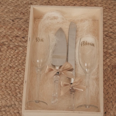 Conjunto de duas taças com nomes, faca e pá de bolo com laços em caixa de madeira sobre tapete de fibra natural